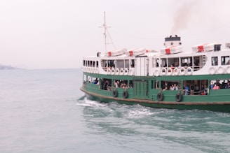 A large passenger ferry is crossing a body of water with several people visible on the deck. The ferry is mainly white and green, with lifebuoys along the side. There is a smokestack emitting smoke, indicating the vessel is powered and in motion. The water is calm, and the sky is overcast.
