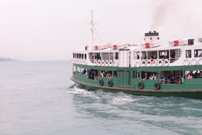 A large passenger ferry is crossing a body of water with several people visible on the deck. The ferry is mainly white and green, with lifebuoys along the side. There is a smokestack emitting smoke, indicating the vessel is powered and in motion. The water is calm, and the sky is overcast.