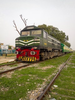 A green and black locomotive with the number 6401 is stationary on the railway tracks. It is connected to a train, and the surrounding area is grassy with scattered stones. A few leafless trees are in the background, along with a railway station sign that reads 'Kharian'.