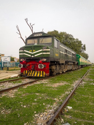A green and black locomotive with the number 6401 is stationary on the railway tracks. It is connected to a train, and the surrounding area is grassy with scattered stones. A few leafless trees are in the background, along with a railway station sign that reads 'Kharian'.