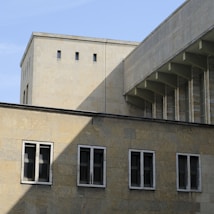 A modern architectural structure with a facade made of light stone tiles. The building features rectangular windows with dark frames and a section of the wall casts a prominent shadow. The sky is clear, providing a minimalistic backdrop to the geometric lines of the architecture.