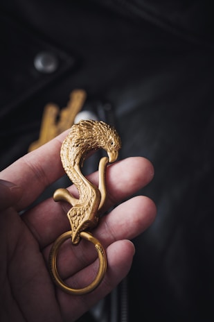 A hand is holding an intricately designed golden keychain shaped like a bird with detailed feather carvings.