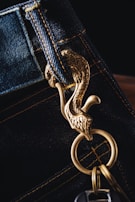 A close-up of a keychain attached to a denim belt loop. The keychain features a decorative gold metal hook shaped like a bird with intricate details. The stitching on the denim is visible, highlighted by yellow thread.