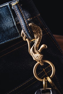 A close-up of a keychain attached to a denim belt loop. The keychain features a decorative gold metal hook shaped like a bird with intricate details. The stitching on the denim is visible, highlighted by yellow thread.