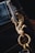 A close-up of a keychain attached to a denim belt loop. The keychain features a decorative gold metal hook shaped like a bird with intricate details. The stitching on the denim is visible, highlighted by yellow thread.