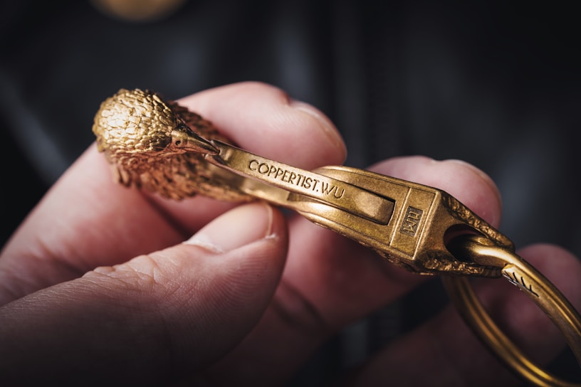 A close-up of a hand holding a metallic object resembling a carabiner, intricately designed to look like a bird with detailed feathers and features. The metal is golden in color and inscribed with the text 'COPPERTIST.WU'. The object's craftsmanship is highlighted by the focus and lighting.