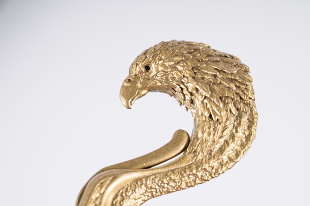 A decorative object featuring an intricately detailed golden eagle head with feathers beautifully textured. It is set against a plain white background, highlighting the metallic sheen and craftsmanship of the object.