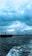 A large cargo ship is sailing on the water under a dramatic, cloudy sky. The dark blue ocean waves appear choppy, indicating windy conditions. In the background, a densely built urban area stretches along the shoreline with multiple buildings visible.