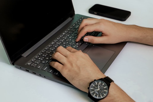 Close-up of hands typing on a laptop keyboard with a smartphone nearby on a desk.