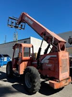 A large orange telescopic forklift labeled 'SkyTrak' is parked on a paved surface near a building. The machinery has big tires and an extended arm with a metal fork attachment. In the background, there is a bright blue sky and a building with white and gray walls. A blue dumpster is also visible beside the forklift.