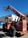A large orange telescopic forklift labeled 'SkyTrak' is parked on a paved surface near a building. The machinery has big tires and an extended arm with a metal fork attachment. In the background, there is a bright blue sky and a building with white and gray walls. A blue dumpster is also visible beside the forklift.