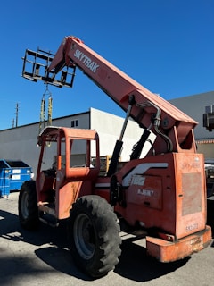 A large orange telescopic forklift labeled 'SkyTrak' is parked on a paved surface near a building. The machinery has big tires and an extended arm with a metal fork attachment. In the background, there is a bright blue sky and a building with white and gray walls. A blue dumpster is also visible beside the forklift.