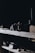 Various old woodworking tools including clamps, chisels, and mallets are placed on wooden shelves in a dimly lit room. The tools are worn and suggest a rustic, vintage setting. Shadows dominate the scene, with a focus on the wood texture and metal parts of the tools.