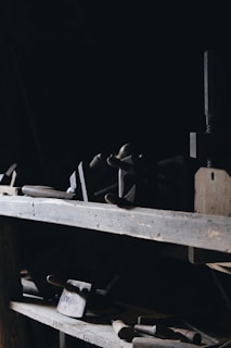 Various old woodworking tools including clamps, chisels, and mallets are placed on wooden shelves in a dimly lit room. The tools are worn and suggest a rustic, vintage setting. Shadows dominate the scene, with a focus on the wood texture and metal parts of the tools.