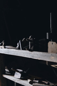 Various old woodworking tools including clamps, chisels, and mallets are placed on wooden shelves in a dimly lit room. The tools are worn and suggest a rustic, vintage setting. Shadows dominate the scene, with a focus on the wood texture and metal parts of the tools.
