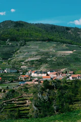 A picturesque village is nestled in a verdant valley surrounded by terraced hills and lush forests. The buildings have traditional European architecture with red roofs and light-colored walls. The landscape is dotted with greenery and appears well-maintained, indicating agricultural activity, possibly vineyards.