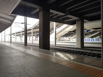 A modern train station platform with empty tracks extending into the distance. The ceiling features large geometric beams, and there are signs displaying Chinese characters. The platform is clean and deserted, with columns evenly spaced along the walkway and bright lighting enhancing the structural details.