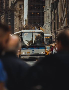 A city street scene with a large bus prominently displayed in the center, marked with 'MTA' and 'X37' heading to 23rd Street FDR Drive. The background shows urban architecture with tall buildings and reflective windows. Yellow taxis and blurred figures in the foreground frame the image.
