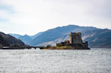 A historic castle situated on a small island surrounded by water, with a stone bridge extending to the mainland. The backdrop consists of rugged mountains under a bright, partly cloudy sky. The area appears serene and untouched, highlighting the scenic beauty of the natural landscape.