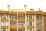 A symmetrical view of apartment buildings with a mustard yellow and cream color scheme. The structures have large windows and balconies with glass railings. Several satellite dishes and antennas are visible on the roof, with some balconies showing curtains and indoor decor.