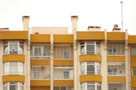 A symmetrical view of apartment buildings with a mustard yellow and cream color scheme. The structures have large windows and balconies with glass railings. Several satellite dishes and antennas are visible on the roof, with some balconies showing curtains and indoor decor.