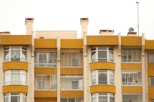 A symmetrical view of apartment buildings with a mustard yellow and cream color scheme. The structures have large windows and balconies with glass railings. Several satellite dishes and antennas are visible on the roof, with some balconies showing curtains and indoor decor.