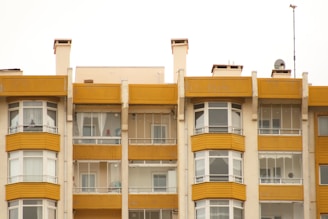 A symmetrical view of apartment buildings with a mustard yellow and cream color scheme. The structures have large windows and balconies with glass railings. Several satellite dishes and antennas are visible on the roof, with some balconies showing curtains and indoor decor.