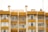 A symmetrical view of apartment buildings with a mustard yellow and cream color scheme. The structures have large windows and balconies with glass railings. Several satellite dishes and antennas are visible on the roof, with some balconies showing curtains and indoor decor.