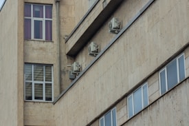 A section of a beige concrete building exterior with multiple windows and several air conditioning units mounted on the walls.