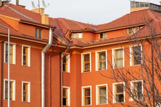 A multi-story building with an orange facade and a red pitched roof. The structure has multiple windows with white frames, some of which are open. A few bare trees are in the foreground, and there are silver metal pipes attached to the building that appear to be part of a drainage or ventilation system. The roof shows some wear and tear, with missing tiles.