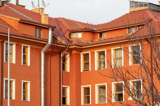 A multi-story building with an orange facade and a red pitched roof. The structure has multiple windows with white frames, some of which are open. A few bare trees are in the foreground, and there are silver metal pipes attached to the building that appear to be part of a drainage or ventilation system. The roof shows some wear and tear, with missing tiles.