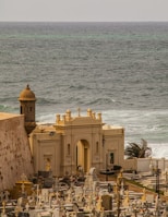 A scenic cemetery by the sea with numerous gravestones and mausoleums. The structure in the foreground features an arched entryway and cross-adorned tops. Waves crash on the rocky shore, accented by distant teal and grey waters. The surrounding landscape includes palm trees buffeted by the wind.