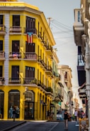 A vibrant street scene features a striking yellow building adorned with a Puerto Rican flag, showcasing its colonial architecture with black wrought-iron balconies. Several people are walking along the bustling sidewalk, which is lined with shops. The sky is clear, and the atmosphere is lively and colorful.