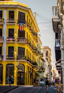 A vibrant street scene features a striking yellow building adorned with a Puerto Rican flag, showcasing its colonial architecture with black wrought-iron balconies. Several people are walking along the bustling sidewalk, which is lined with shops. The sky is clear, and the atmosphere is lively and colorful.
