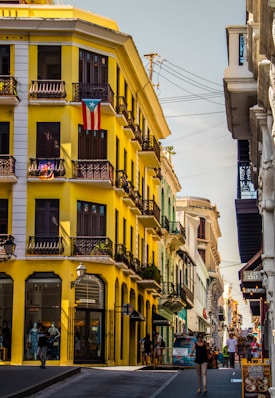 A vibrant street scene features a striking yellow building adorned with a Puerto Rican flag, showcasing its colonial architecture with black wrought-iron balconies. Several people are walking along the bustling sidewalk, which is lined with shops. The sky is clear, and the atmosphere is lively and colorful.