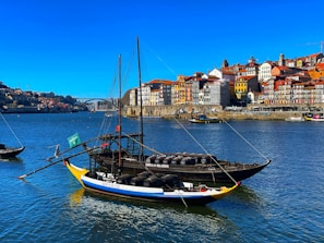 A scenic riverside view features a traditional brightly painted boat on a calm river with barrels on board, likely used for transporting wine. A row of colorful buildings lines the waterfront, with historical architecture visible in the background. A bridge spans the river in the distance under a clear blue sky.