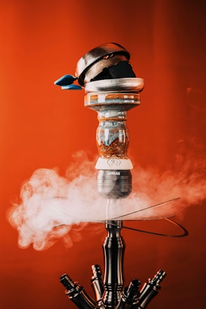 A detailed close-up of a hookah with smoke emanating from it. The hookah has a modern and metallic design with a decorative bowl containing coal on top. The background is a solid, vibrant red color, enhancing the contrast of the metallic elements and the white smoke.