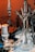 A close-up of an elaborate hookah setup featuring a clear glass base with metal components. There are multiple hoses and attachments, along with a suspended accessory. Various other objects, including metallic and plastic components, are scattered around on a patterned surface.