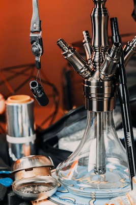 A close-up of an elaborate hookah setup featuring a clear glass base with metal components. There are multiple hoses and attachments, along with a suspended accessory. Various other objects, including metallic and plastic components, are scattered around on a patterned surface.