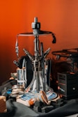 A detailed view of a hookah set on a table, surrounded by related accessories such as charcoal cubes and a charcoal burner. The hookah features a transparent glass base with multiple hose connectors, and it sits on a decorative round plate. The background features a warm, orange-toned setting, creating an inviting atmosphere with various items like a leather pouch visible around the setup.