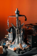 A detailed view of a hookah set on a table, surrounded by related accessories such as charcoal cubes and a charcoal burner. The hookah features a transparent glass base with multiple hose connectors, and it sits on a decorative round plate. The background features a warm, orange-toned setting, creating an inviting atmosphere with various items like a leather pouch visible around the setup.