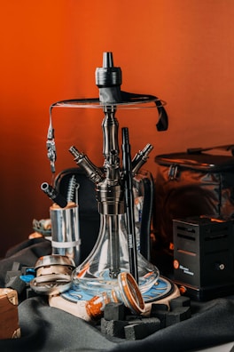 A detailed view of a hookah set on a table, surrounded by related accessories such as charcoal cubes and a charcoal burner. The hookah features a transparent glass base with multiple hose connectors, and it sits on a decorative round plate. The background features a warm, orange-toned setting, creating an inviting atmosphere with various items like a leather pouch visible around the setup.