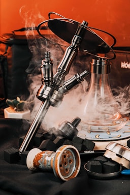 A collection of hookah components featuring a metallic hookah stem with a black tray on top, surrounded by smoke. Nearby are various accessories including charcoal cubes, a ceramic bowl, and a glass base. The background has a warm orange hue, enhancing the contrast against the dark surface.