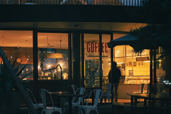 A cozy coffee shop with glowing warm lights seen through large windows. The interior is dimly lit with various light fixtures and a person standing at the counter. Empty metal chairs and tables are arranged outside under an umbrella. Various decorations and plants enhance the inviting atmosphere inside.