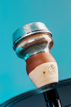 A close-up of a hookah bowl, featuring a mix of metal and ceramic materials. The upper part is metallic, while the lower part has a textured, brown ceramic base. The background is a solid teal color.