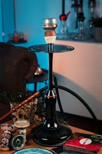 A hookah setup placed on a table, featuring a large black stem and hose with a clay bowl on top. Surrounding the hookah are various accessories, including replacement bowls and a package of aluminum foil. The scene is warmly lit, with some green plant leaves as part of the decor. The background includes other hookahs on display.