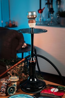 A hookah setup placed on a table, featuring a large black stem and hose with a clay bowl on top. Surrounding the hookah are various accessories, including replacement bowls and a package of aluminum foil. The scene is warmly lit, with some green plant leaves as part of the decor. The background includes other hookahs on display.