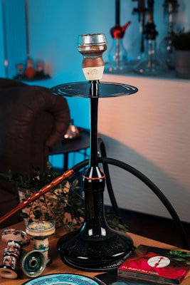 A hookah setup placed on a table, featuring a large black stem and hose with a clay bowl on top. Surrounding the hookah are various accessories, including replacement bowls and a package of aluminum foil. The scene is warmly lit, with some green plant leaves as part of the decor. The background includes other hookahs on display.