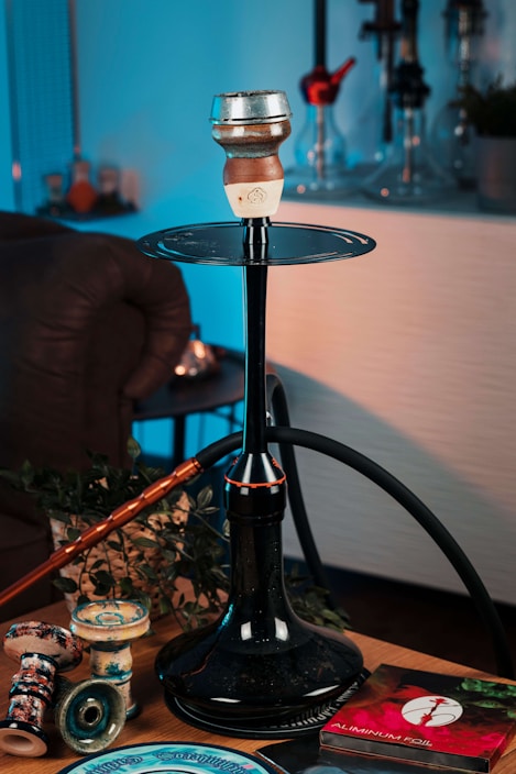 A hookah setup placed on a table, featuring a large black stem and hose with a clay bowl on top. Surrounding the hookah are various accessories, including replacement bowls and a package of aluminum foil. The scene is warmly lit, with some green plant leaves as part of the decor. The background includes other hookahs on display.