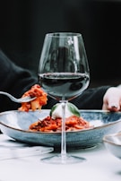 A glass of red wine is set in front of a plate of pasta with a rich tomato sauce and a fresh basil leaf. A person is holding a fork with a portion of pasta above the plate, and another hand is seen resting near the plate.